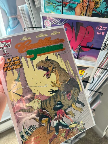 The same hand at the same spinner rack, but this time holding the aforementioned Cadillacs & Dinosaurs issue, which depicts a male and female-presenting protagonists facing off against two huge tyrannosaurus-like monsters in an arid wasteland. The logo is shiny!