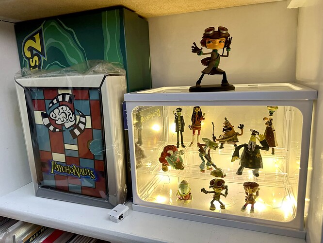 A white shelf appointed with collector's edition copies of Psychonauts 1 and 2, a PS4 copy of Psychonauts 2, a large figure of Raz, and an illuminated display containing ten blind-box Psychonauts figurines